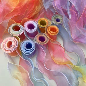 Könnte beinhalten: Eine Sammlung mehrerer Rollen aus durchsichtigem, transparentem Organzaband in einem lebendigen Farbverlauf aus Orange, Pink, Lila und Blau. Jede Rolle hat einen gewellten, gerüschten Rand mit einer feinen Verzierung aus goldenem Faden.