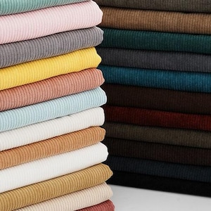 May include: Stacks of colorful corduroy fabric in various shades, including teal, pink, gray, yellow, and brown. The fabric has a ribbed texture and is neatly folded, showcasing a range of colors suitable for apparel or home decor projects.