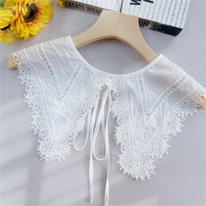 Puede incluir: Un cuello de encaje blanco con un delicado dise&ntilde;o floral y cierre de lazo. El cuello est&aacute; hecho de una tela ligera y presenta intrincados detalles de encaje a lo largo de los bordes. El cuello se muestra en una percha de madera.