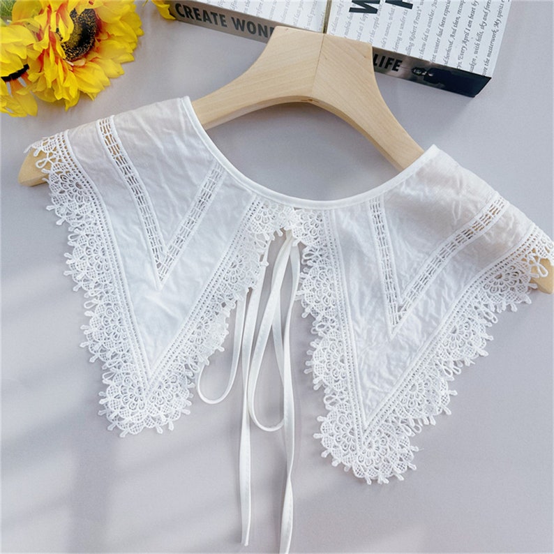 Puede incluir: Un cuello de encaje blanco con un delicado dise&ntilde;o floral y cierre de lazo. El cuello est&aacute; hecho de una tela ligera y presenta intrincados detalles de encaje a lo largo de los bordes. El cuello se muestra en una percha de madera.