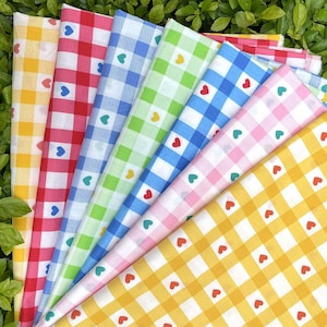 May include: A collection of colorful gingham fabric swatches. Each swatch features a different color, including yellow, red, blue, and green, with a white grid pattern. Small heart-shaped accents are scattered across the fabric.