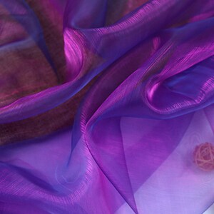 May include: Purple and blue iridescent sheer fabric with a slight shimmer. The fabric is draped and folded, creating soft folds and waves.