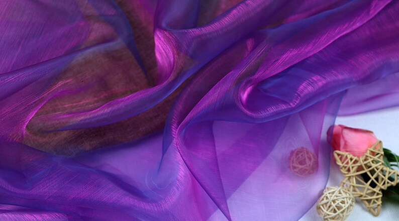 May include: Purple and blue iridescent sheer fabric with a slight shimmer. The fabric is draped and folded, creating soft folds and waves.