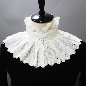 May include: A white lace collar with a ruffled design. The collar features intricate cutwork and a button closure. The collar is displayed on a mannequin, set against a black background.