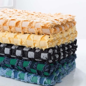 Peut inclure: Une pile de quatre carrés de tissu texturé à franges, dans les tons de jaune, noir et blanc, vert et bleu. Le tissu présente un motif tissé à carreaux. La pile est soigneusement disposée, mettant en valeur les différentes couleurs et textures.
