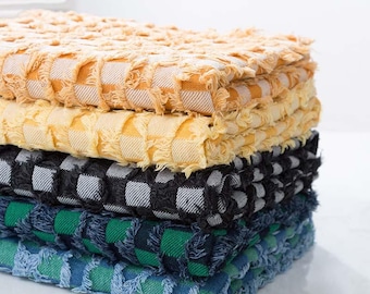 May include: A stack of four textured fabric squares with fringed edges, in yellow, black and white, green, and blue. The fabric has a woven, checkered pattern. The stack is neatly arranged, showcasing the different colours and textures.