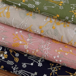 May include: Close-up of folded fabric in shades of green, tan, pink, and navy blue. Each fabric is embroidered with a delicate floral pattern in white and yellow. The fabrics are stacked on a wooden surface.
