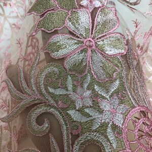 May include: Close-up of a sheer fabric with intricate floral embroidery. The design features large flowers with white and pink petals, outlined in pink thread, and gold-colored leaves. The background is a soft, neutral tone.