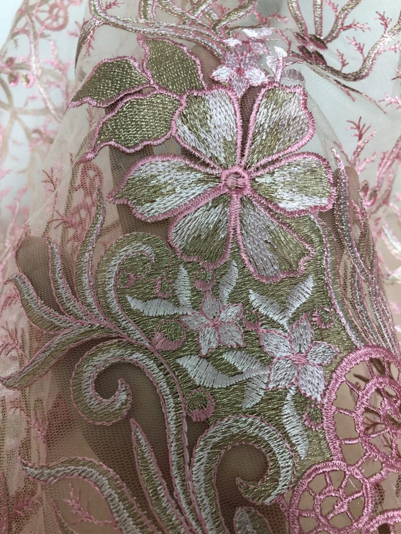 May include: Close-up of a sheer fabric with intricate floral embroidery. The design features large flowers with white and pink petals, outlined in pink thread, and gold-colored leaves. The background is a soft, neutral tone.