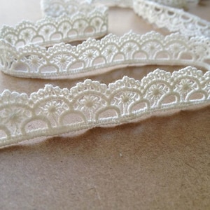 May include: Close-up of ivory-colored lace trim with a scalloped edge. The trim features a repeating pattern of semi-circles and floral details. The lace is soft and delicate, perfect for sewing and craft projects.