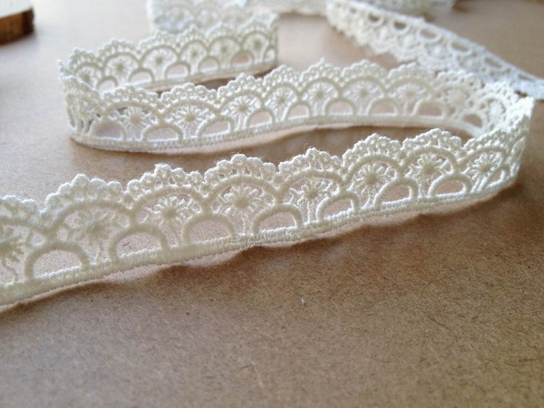 May include: Close-up of ivory-colored lace trim with a scalloped edge. The trim features a repeating pattern of semi-circles and floral details. The lace is soft and delicate, perfect for sewing and craft projects.