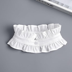 Puede incluir: Un cuello blanco con volantes y cierre de botón. El cuello está hecho de una tela suave y presenta un diseño plisado en capas. El cuello es un accesorio de moda.
