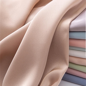 May include: Close-up of flowing fabric in a soft, muted peach colour. The fabric has a subtle sheen and a delicate texture. A stack of various pastel-coloured fabrics is visible in the background, including lavender, pink, and blue.