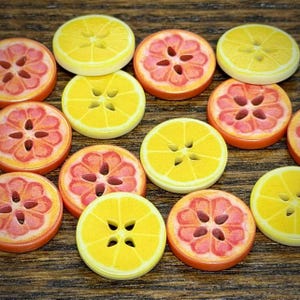 Puede incluir: Una colección de botones redondos con forma de rodajas de cítricos, con diseños de limones amarillos y pomelos rosas. Cada botón tiene cuatro orificios centrales dispuestos en forma cuadrada, con un acabado liso y detalles de segmentos.