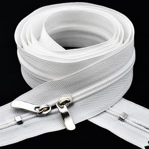 Puede incluir: Cremallera blanca de doble cara con tiradores de metal plateado, enrollada sobre un fondo negro. Los dientes de la cremallera son visibles y la tela tiene un sutil patrón de espiga. Ideal para proyectos de costura.