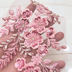 Puede incluir: Primer plano de una aplicación floral rosa. El diseño presenta múltiples flores de tela rosa, adornadas con lentejuelas, cuentas y cristales. Delicadas hojas y cuentas con forma de perla también están incorporadas en el diseño, todo sobre un fondo de malla transparente.