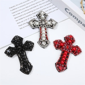 Pode incluir: Três enfeites decorativos em forma de cruz. Um é preto, outro vermelho e o outro é uma mistura de branco, prata e vermelho. Cada cruz é adornada com contas e cristais, criando um efeito brilhante. As cruzes estão em uma superfície branca.