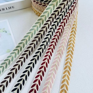 Könnte beinhalten: Nahaufnahme mehrerer Spulen dekorativen Bandes mit einem Blattmuster. Die Bänder sind in verschiedenen Farben erhältlich, darunter Grün, Braun, Schwarz, Rot, Rosa und Gelb, mit goldenen Akzenten. Die Bänder sind diagonal angeordnet und präsentieren ihr Design.
