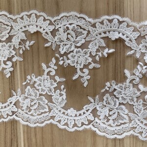 May include: White floral lace trim with a scalloped edge. The lace features detailed floral and leaf patterns. The trim is ideal for sewing, crafting, or adding a delicate touch to garments or home decor projects.