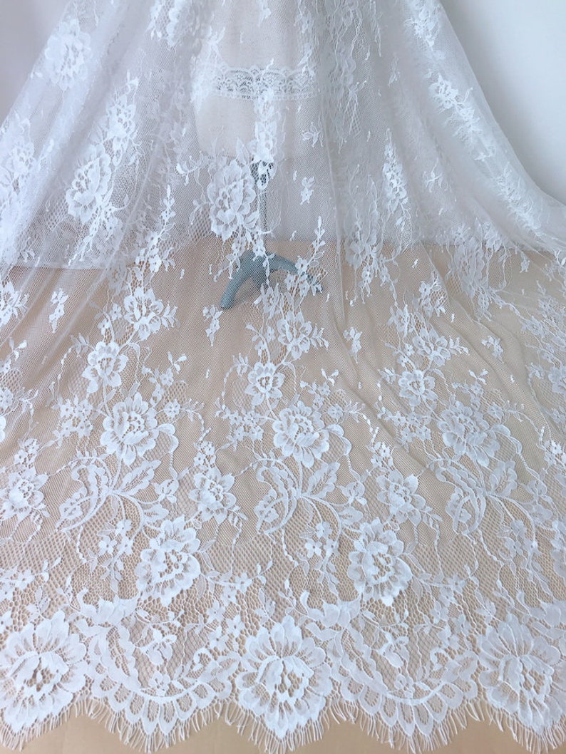 May include: White floral lace fabric with a delicate pattern. The lace has a scalloped edge and is sheer.