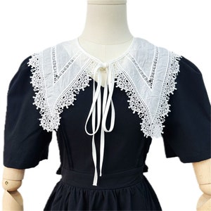 Puede incluir: Vestido negro con cuello blanco ribeteado con encaje. El cuello tiene un dise&ntilde;o puntiagudo y est&aacute; atado con cintas blancas. El vestido tiene mangas cortas y una cintura entallada, creando un look cl&aacute;sico y elegante.