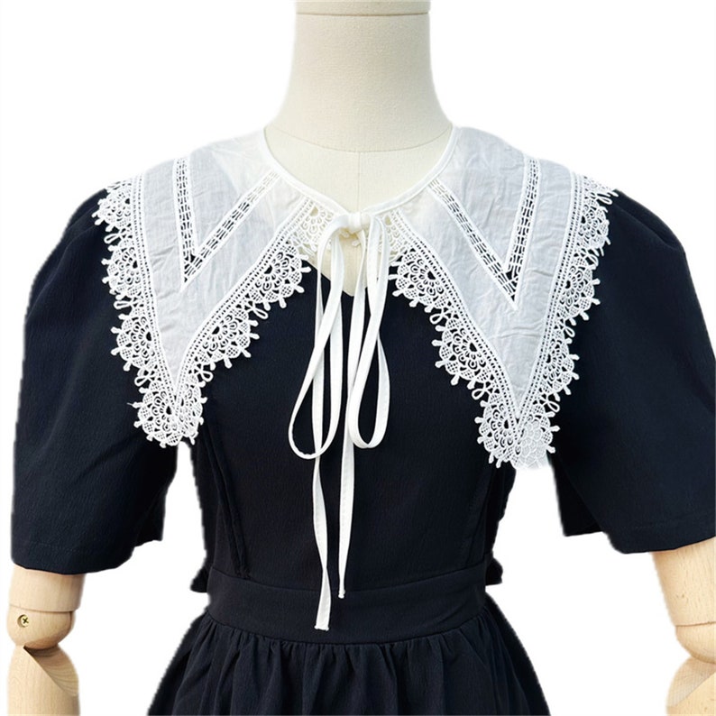 Puede incluir: Vestido negro con cuello blanco ribeteado con encaje. El cuello tiene un dise&ntilde;o puntiagudo y est&aacute; atado con cintas blancas. El vestido tiene mangas cortas y una cintura entallada, creando un look cl&aacute;sico y elegante.