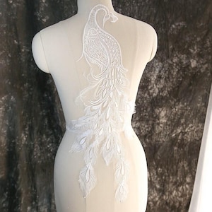 May include: A white lace applique featuring a detailed peacock design with intricate embroidery and feather patterns. The decorative piece is displayed on a mannequin torso, showcasing the elaborate stitching and the long, flowing tail feathers.