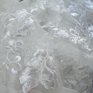 May include: White tulle fabric with delicate floral embroidery and sequins. The fabric has a soft, flowing texture and is perfect for creating elegant garments or accessories.