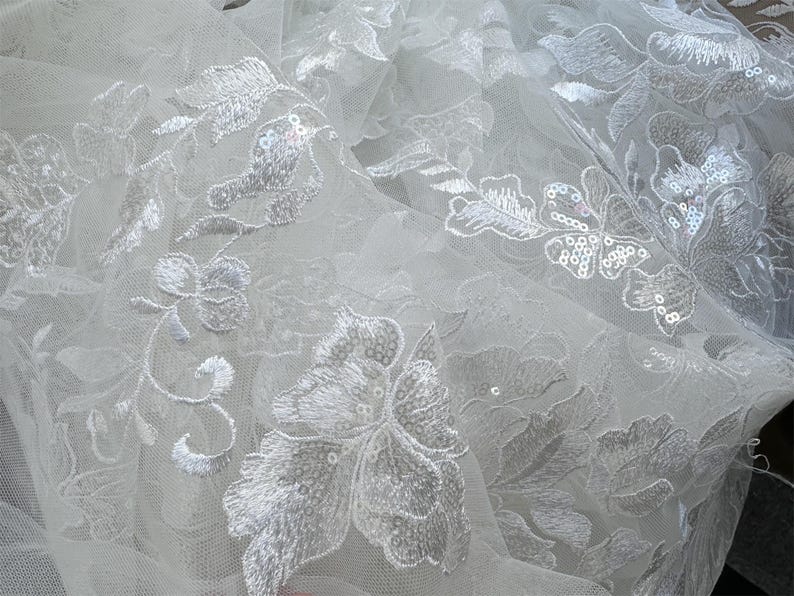 May include: White tulle fabric with delicate floral embroidery and sequins. The fabric has a soft, flowing texture and is perfect for creating elegant garments or accessories.