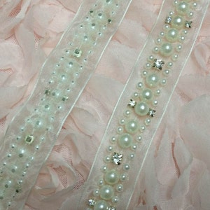 May include: Two white beaded trims with pearl-like beads and clear rhinestones on a pink fabric background.