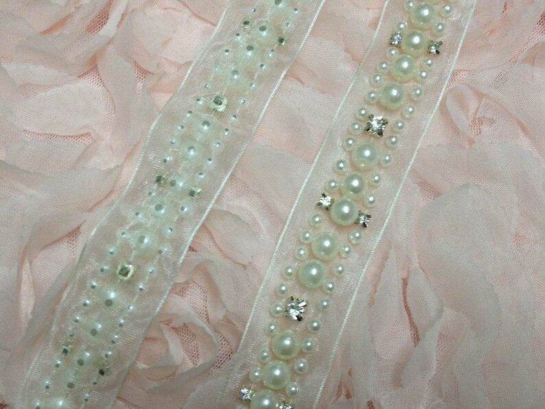 May include: Two white beaded trims with pearl-like beads and clear rhinestones on a pink fabric background.