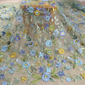Pode incluir: Tecido transparente com bordados florais. A borda apresenta flores azuis, amarelas e roxas com folhas e caules verdes. O tecido é de cor clara e neutra, com flores detalhadas.