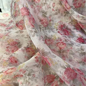 May include: Sheer fabric with a floral pattern featuring pink roses, yellow accents, and gray leaves on a white background. The fabric is lightweight and appears to be flowing, suitable for apparel or home decor projects.