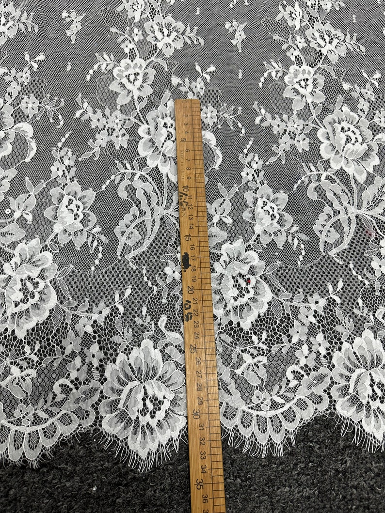 May include: White floral lace fabric with a delicate pattern. The fabric is shown next to a ruler measuring 36 inches.