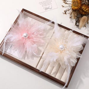 May include: Two decorative floral hair accessories. One is light pink, the other is cream-coloured, both with a large pearl centre, surrounded by feathers and small beads. They are displayed in a wooden jewellery box.