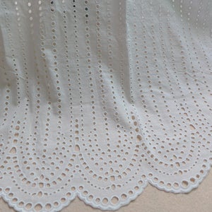 May include: White cotton fabric with a delicate eyelet pattern. The fabric has a scalloped edge and is ideal for crafting and sewing projects.