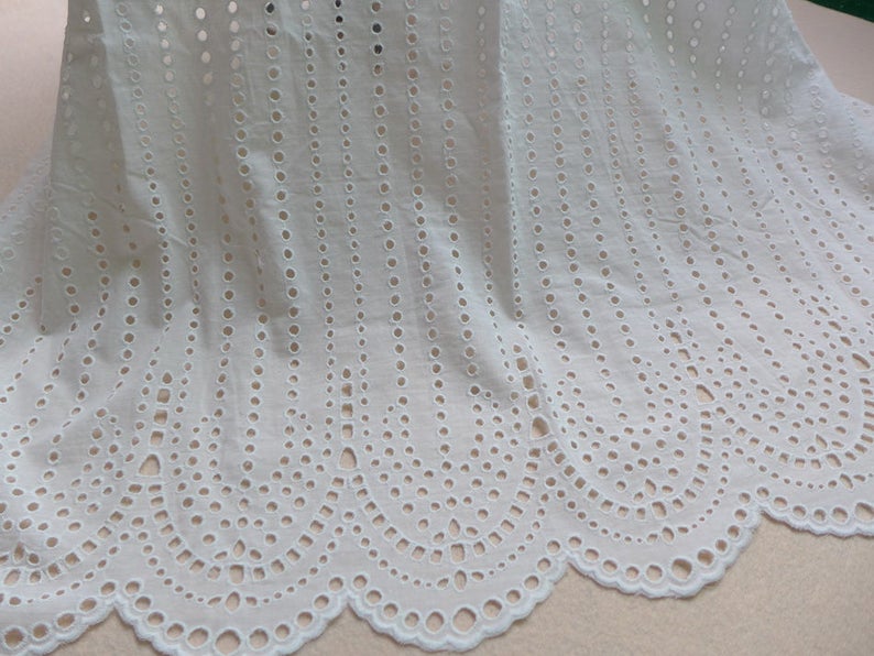 May include: White cotton fabric with a delicate eyelet pattern. The fabric has a scalloped edge and is ideal for crafting and sewing projects.