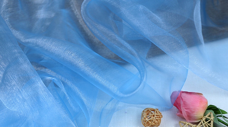May include: Light blue sheer fabric with a soft, flowing texture. The fabric is draped over a white surface.