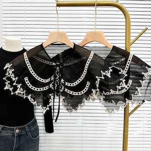 以下が含まれることがあります： 木製のハンガーに掛けられた、白い刺繍トリムが付いた2つの黒いシアーレースカラー。襟はスカラップエッジとタイクロージャーを備えています。襟は金の衣類ラックに吊るされています。