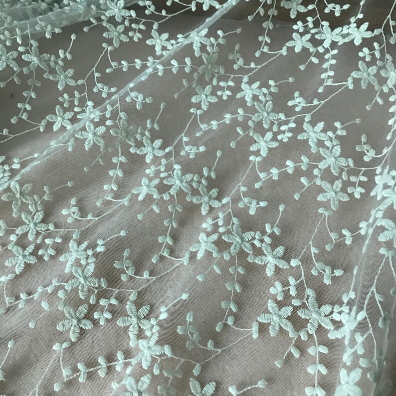 K&ouml;nnte beinhalten: Blassgr&uuml;ne florale Stickerei auf einem durchsichtigen wei&szlig;en Stoff. Das Design zeigt zarte Blumen und Bl&auml;tter.