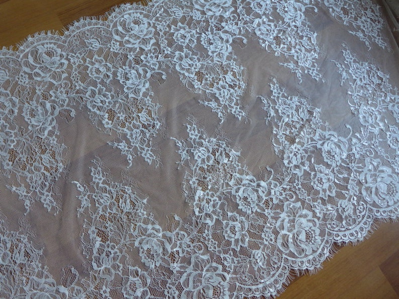 May include: White lace fabric with a delicate floral pattern. The lace has a scalloped edge and is sheer.