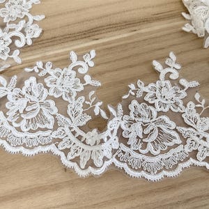 May include: Close-up of white floral lace trim with a scalloped edge. The delicate lace features detailed embroidered flowers and leaves. The lace is set against a light brown wooden surface, highlighting the intricate design.