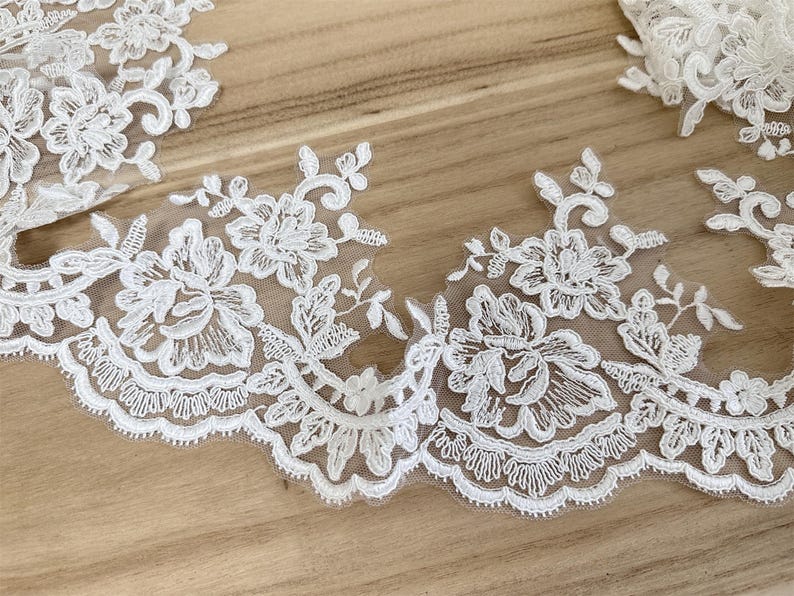May include: Close-up of white floral lace trim with a scalloped edge. The delicate lace features detailed embroidered flowers and leaves. The lace is set against a light brown wooden surface, highlighting the intricate design.