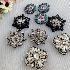 Może przedstawiać: Asortyment ozdób dekoracyjnych, w tym wzory w kształcie gwiazd i kwiatów. Ozdoby mają różne kolory, w tym czarny, srebrny i opalizujące odcienie klejnotów. Niektóre mają perłowe akcenty. Prawdopodobnie są przeznaczone do odzieży lub projektów rzemieślniczych.