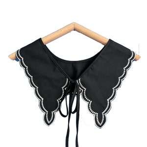 May include: A black fabric collar with a scalloped edge and a white pearl trim. The collar has a tie at the back.