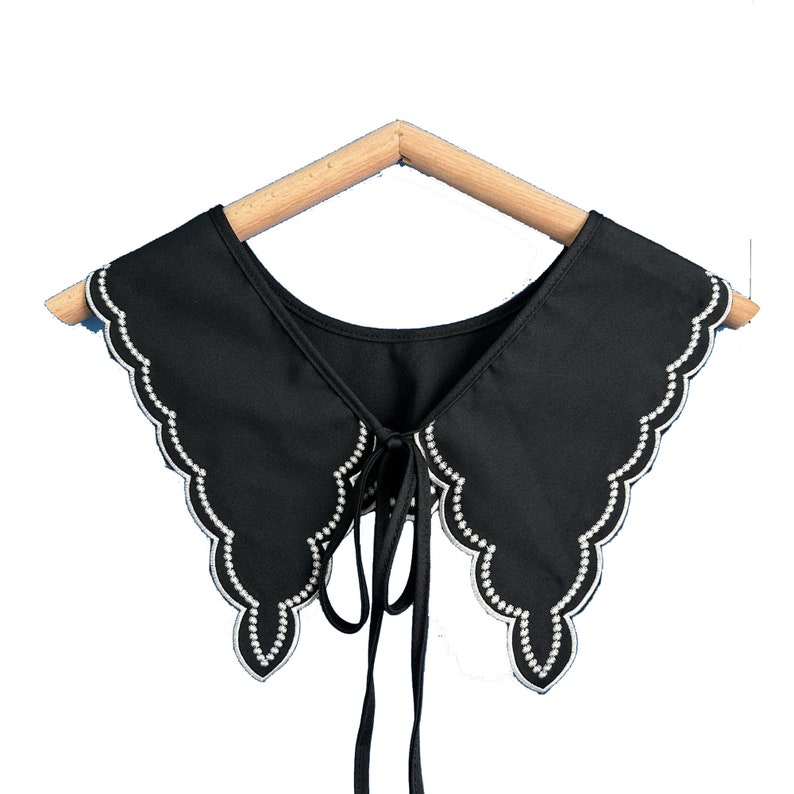 May include: A black fabric collar with a scalloped edge and a white pearl trim. The collar has a tie at the back.