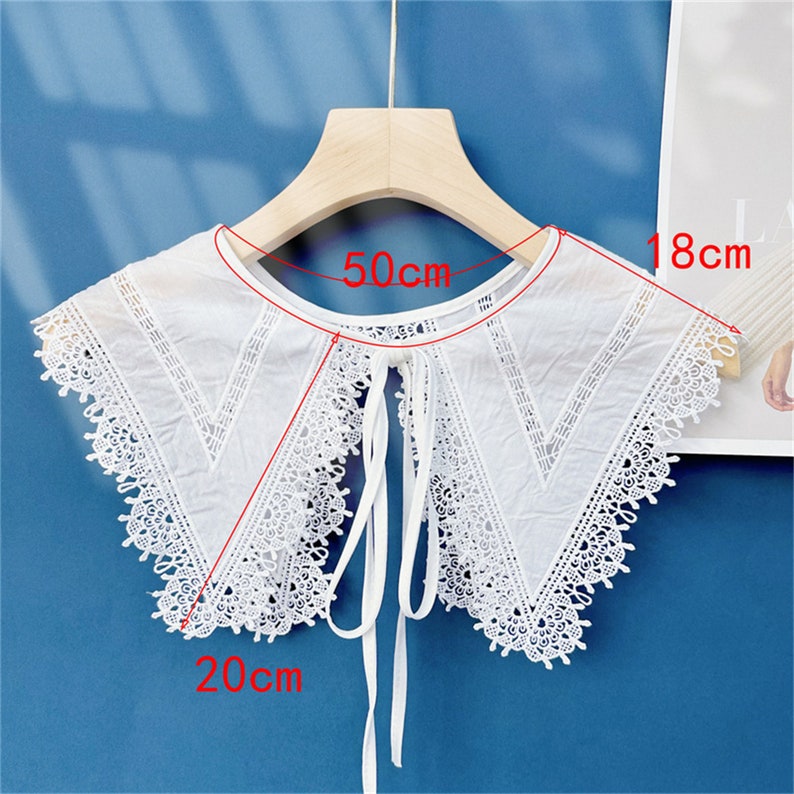 Puede incluir: Un cuello de encaje blanco, de 20 cm de ancho y 18 cm de alto, con un escote de 50 cm, cuelga de una percha de madera. El cuello presenta un delicado ribete de encaje y un cierre de lazo.