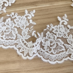 May include: Close-up of white floral lace trim with scalloped edges. The lace features detailed embroidered flowers and leaves on a sheer background. The trim is ideal for sewing and craft projects.