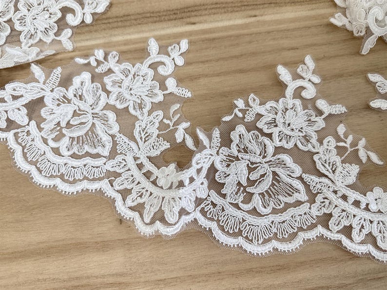 May include: Close-up of white floral lace trim with scalloped edges. The lace features detailed embroidered flowers and leaves on a sheer background. The trim is ideal for sewing and craft projects.