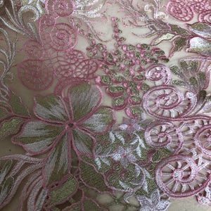 May include: Close-up of embroidered fabric featuring a floral design. The fabric has a sheer, light beige background with intricate embroidery in pink, gold, and white. The design includes flowers, swirls, and other decorative elements.